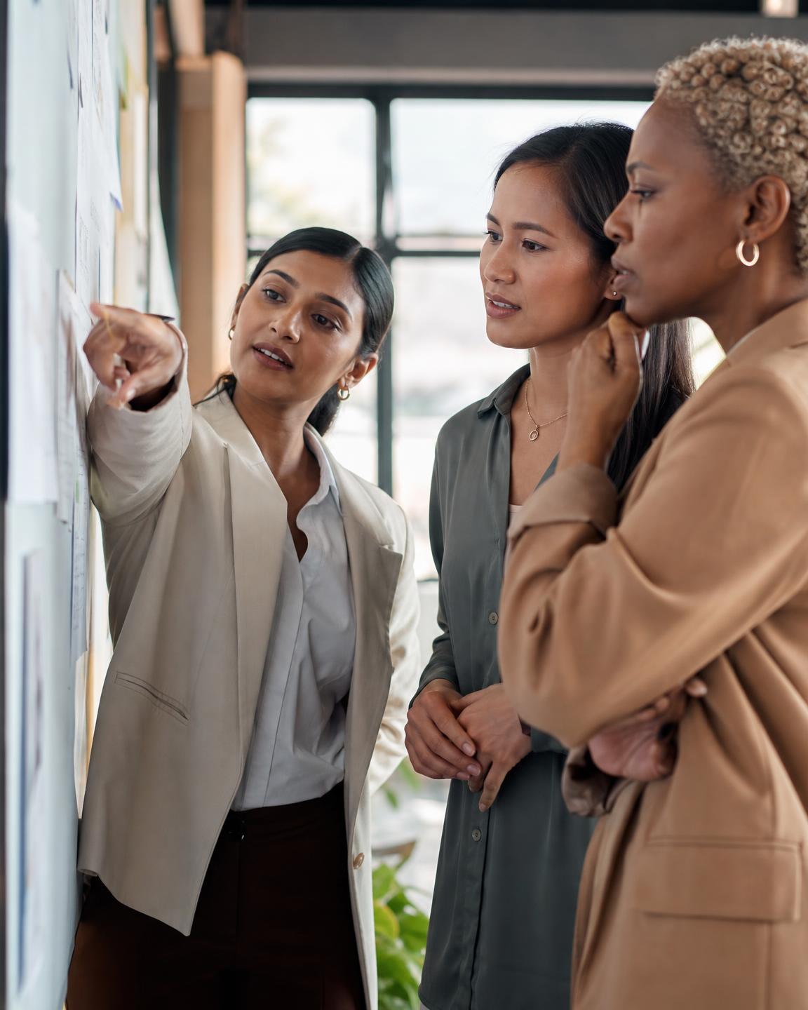 Three diverse businesswomen reviewing a strategic plan together on a wall board in a modern office